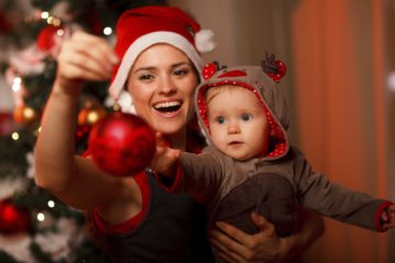 Happy mother showing Christmas ball  to baby near Christmas tree