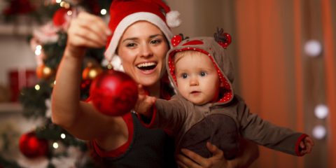 Happy mother showing Christmas ball  to baby near Christmas tree