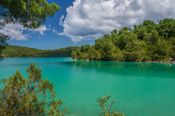 Clean, clear turquoise green lake water surrounded by trees and Mediterranean vegetation. Blue sky with beautiful white clouds.
