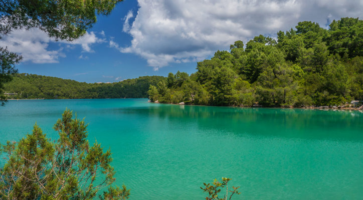 Clean, clear turquoise green lake water surrounded by trees and Mediterranean vegetation. Blue sky with beautiful white clouds.